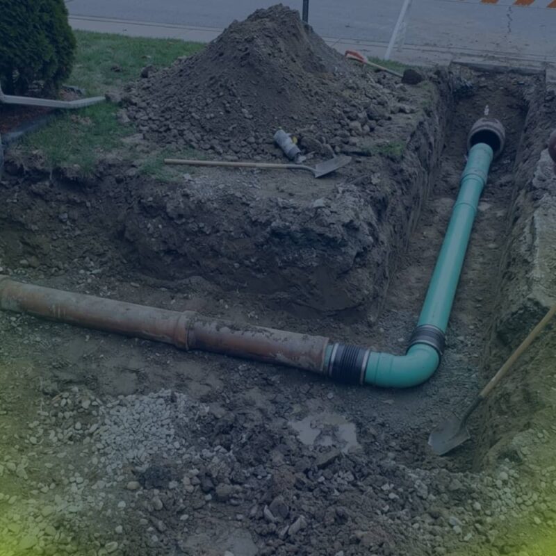 A construction worker stands in an excavated trench where a new green PVC sewer lateral pipe is being connected to an existing iron line leading from a residential foundation.
