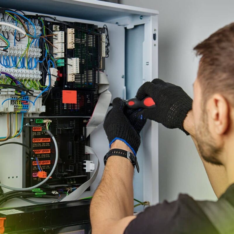 A technician wearing black gloves uses a screwdriver to work on a complex electrical control panel filled with multi-colored wiring and circuit boards.