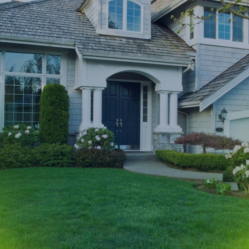 A multi-story suburban home featuring gray shingle siding, a dark blue front door, and a beautifully manicured green lawn with landscaped bushes.