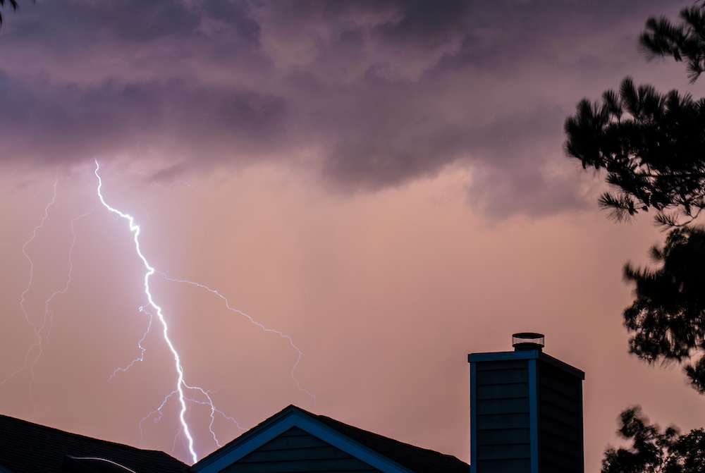 A bolt of lightning strikes near a house roof during a severe thunderstorm, illustrating how atmospheric pressure changes from storms can raise indoor radon levels.